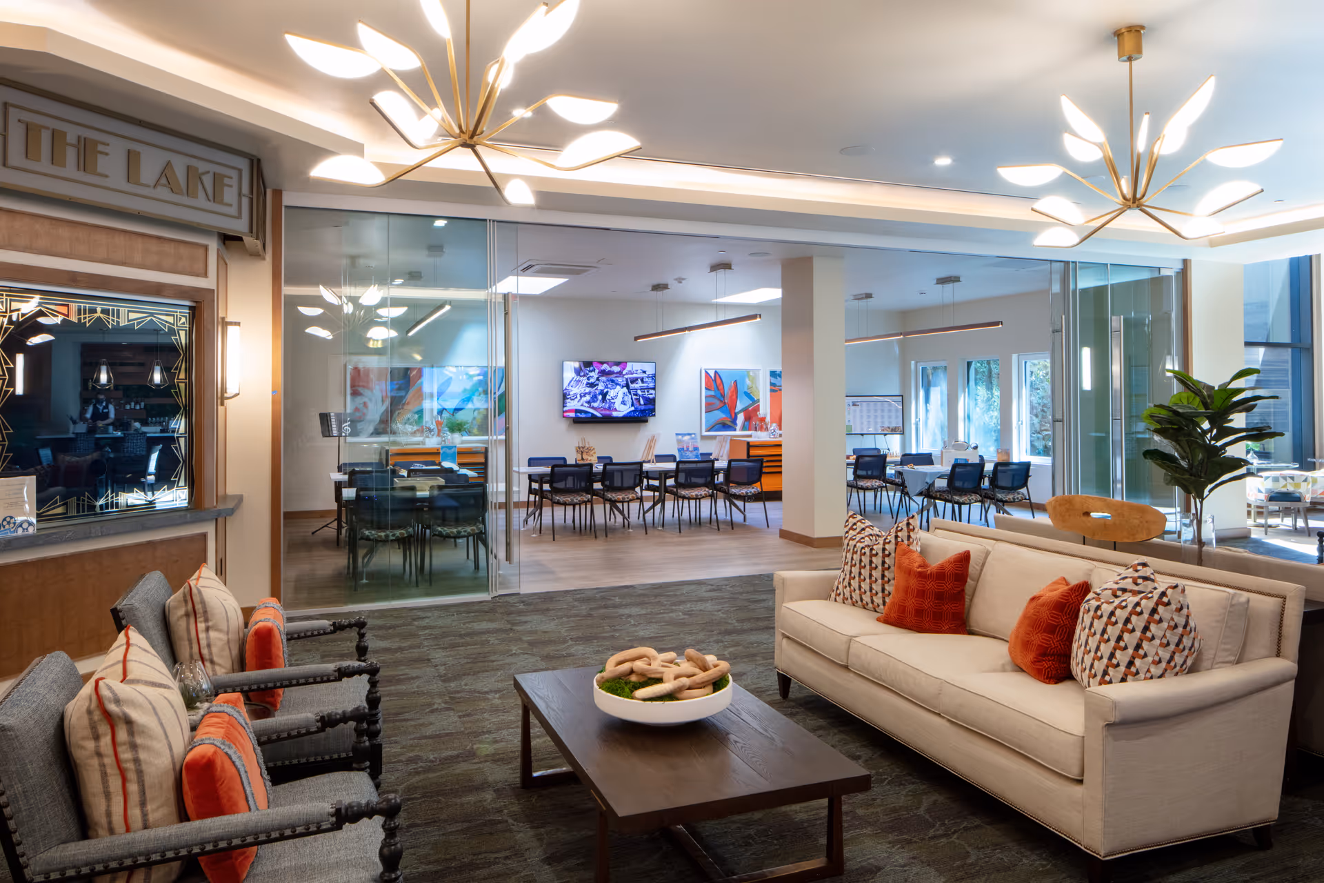 A modern and well-lit common area in a senior living facility featuring a beige sofa with patterned and orange pillows, two armchairs with striped and orange cushions, a wooden coffee table with a decorative bowl, and a glass-walled meeting or dining room in the background with tables and chairs. The space has contemporary light fixtures and a plant near the window.