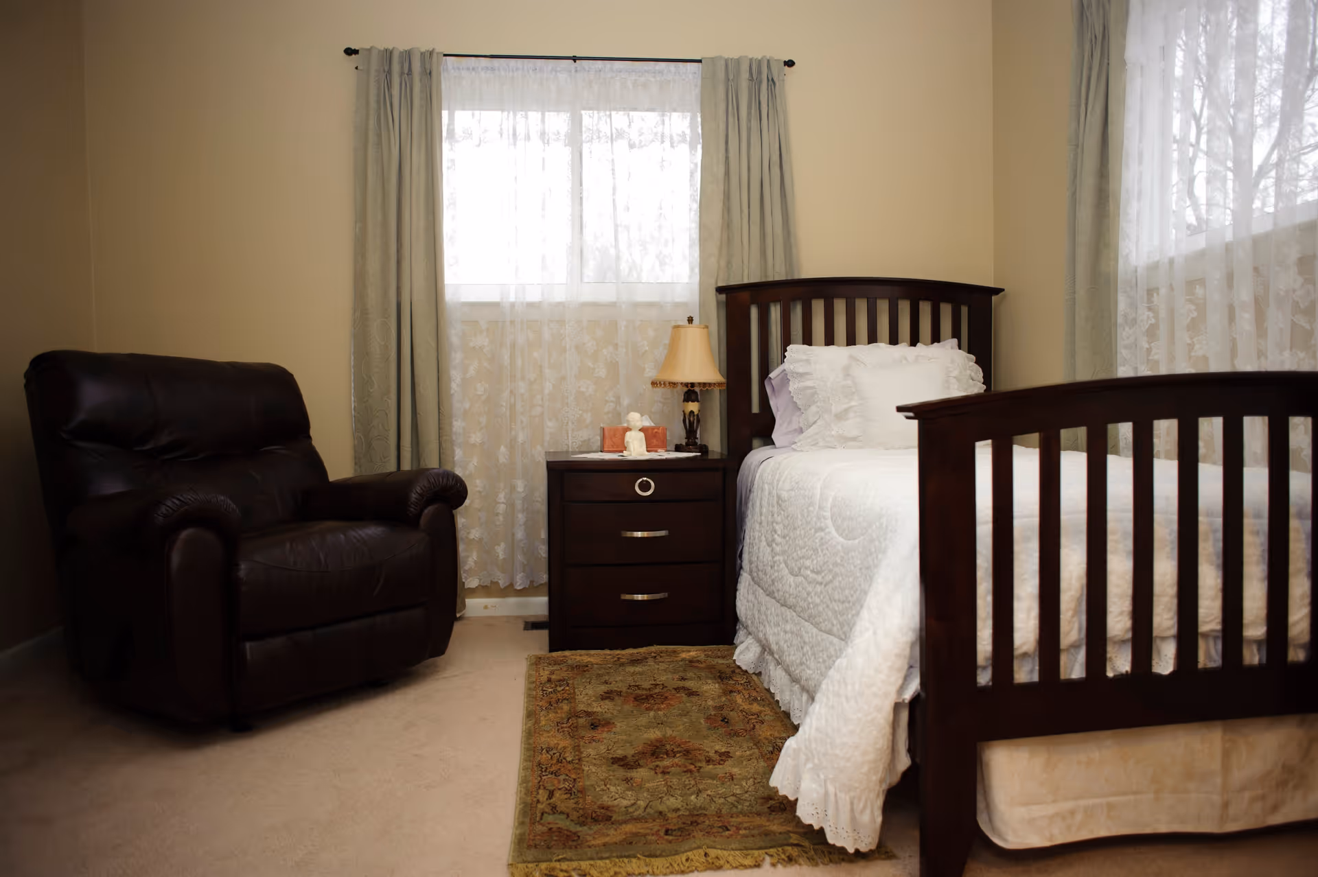 A cozy bedroom with a dark wooden bed frame and white bedding, a matching dark wooden nightstand with a lamp and decorative items, a dark brown leather armchair, beige walls, carpeted floor, two windows with sheer white curtains and light green drapes, and a small patterned rug beside the bed.