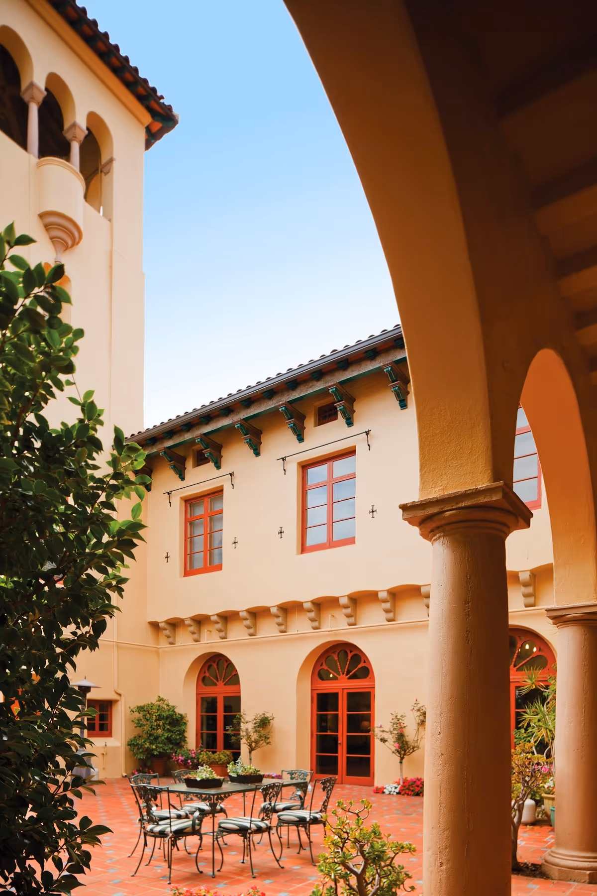 Outdoor courtyard area at Casa Dorinda featuring a round metal table with six cushioned chairs on a terracotta tiled floor, surrounded by potted plants and trees. The courtyard is enclosed by a beige building with red-framed windows and arched doorways, visible under a clear blue sky.