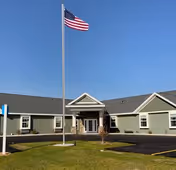 Exterior view of a single-story assisted living facility building with a gray roof and light green siding. A flagpole with an American flag is prominently displayed in front of the building under a clear blue sky. The entrance has a small covered porch with stone accents on the lower walls.