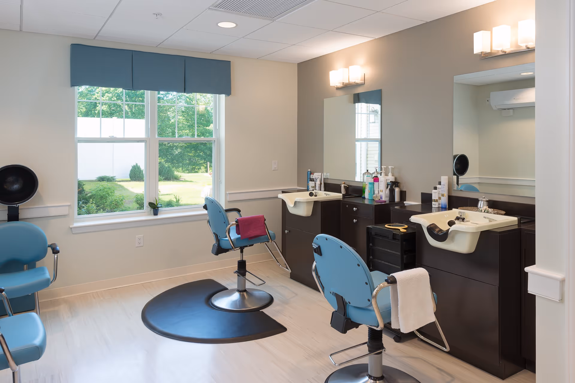 Interior of a hair salon area with two blue salon chairs in front of sinks and mirrors. The room has light-colored walls, a large window with a blue valance, and various hair care products on the counter.