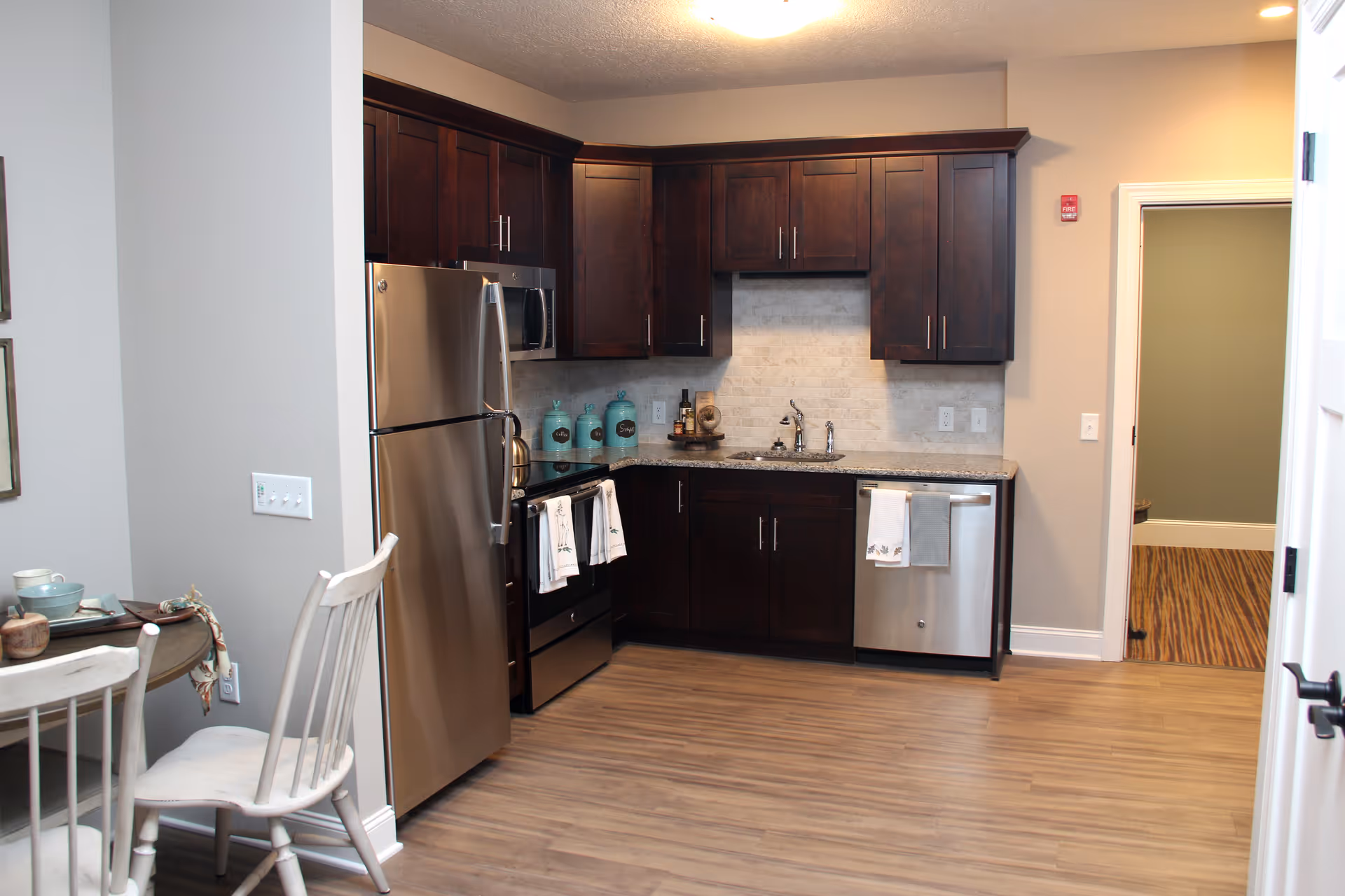 Small modern kitchen with dark wood cabinets, stainless steel appliances, and a small dining table to the left.