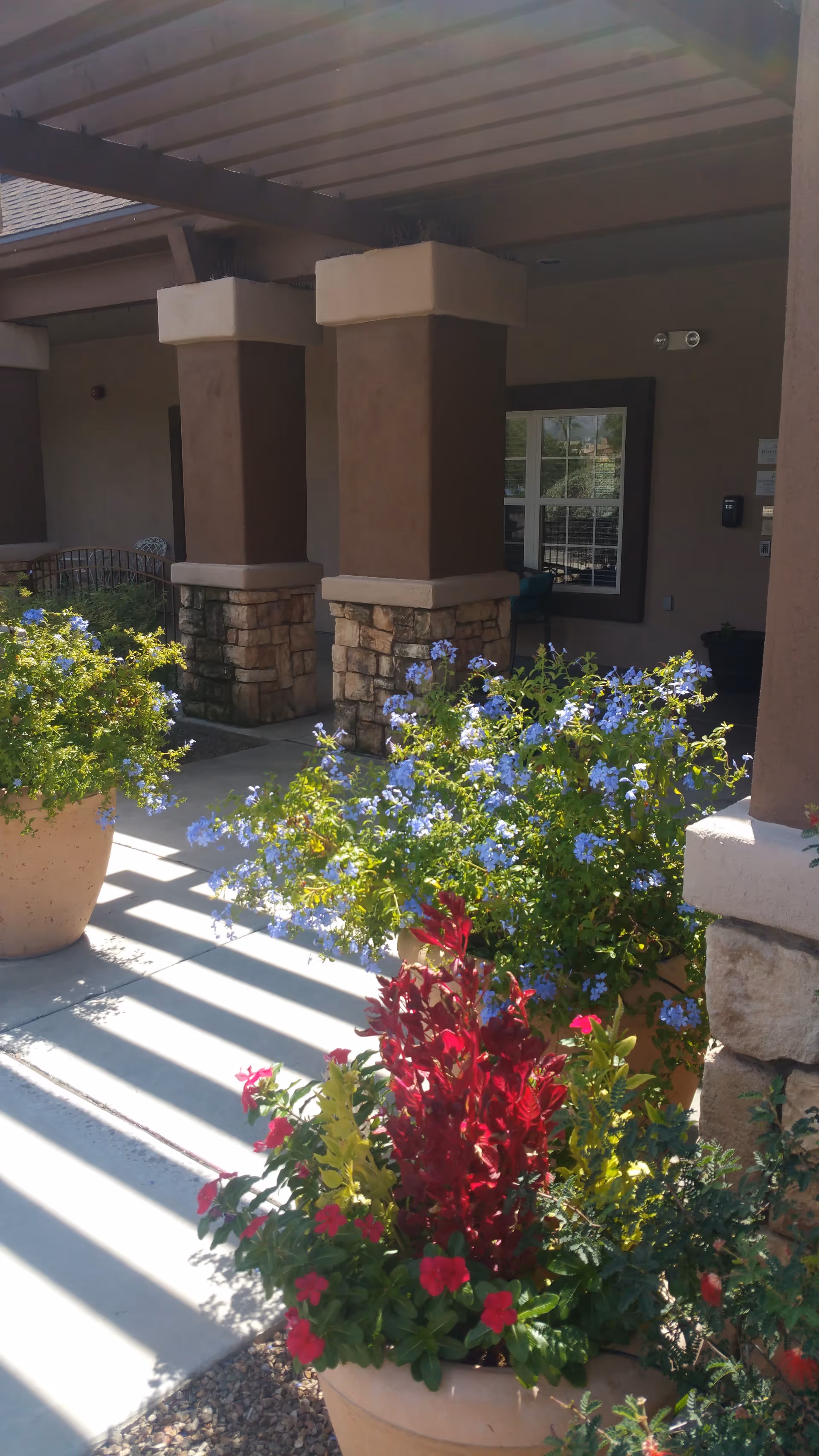 Covered building entrance with stone columns and flowering planters along a sunny walkway