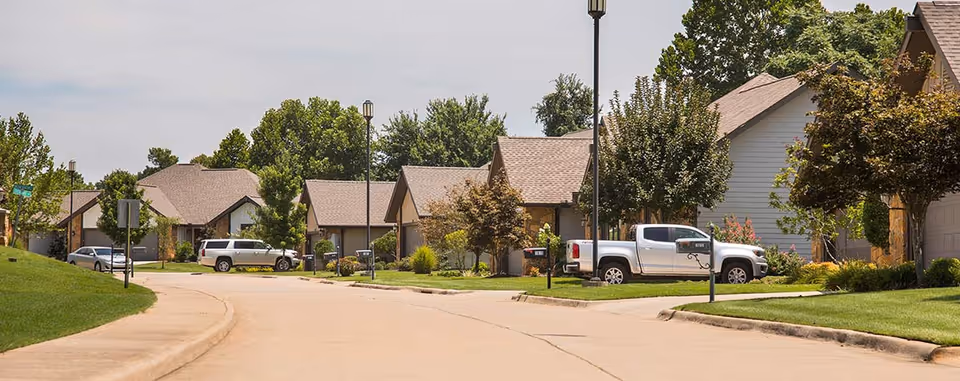 A quiet residential street in a senior living community with single-story houses, well-maintained lawns, trees, and parked vehicles including a white pickup truck and an SUV.