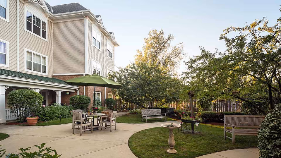 Outdoor patio area at a senior living facility with a round table and four chairs under a green umbrella, surrounded by bushes, trees, and benches. The building exterior is visible with multiple windows and beige siding.