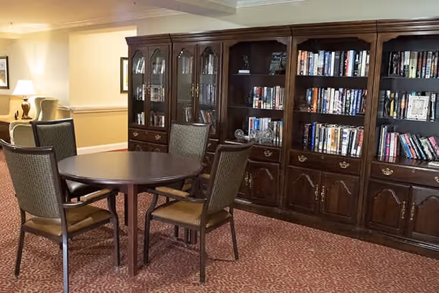 A cozy interior room with a round wooden table surrounded by four chairs. Behind the table is a large wooden bookshelf filled with books and decorative items. The room has warm lighting and carpeted flooring with a patterned design.