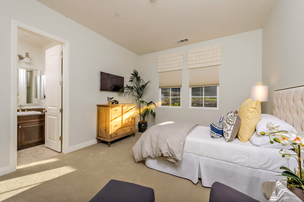Well-lit bedroom with a made bed, bedside lamp, dresser and wall-mounted TV, with an open door showing an ensuite bathroom.