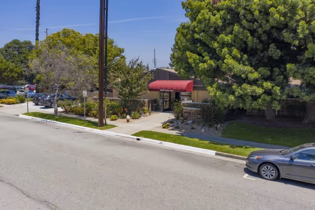 Exterior view of a single-story building with a red awning over the entrance, surrounded by trees and landscaping, with a parked car on the street in front.