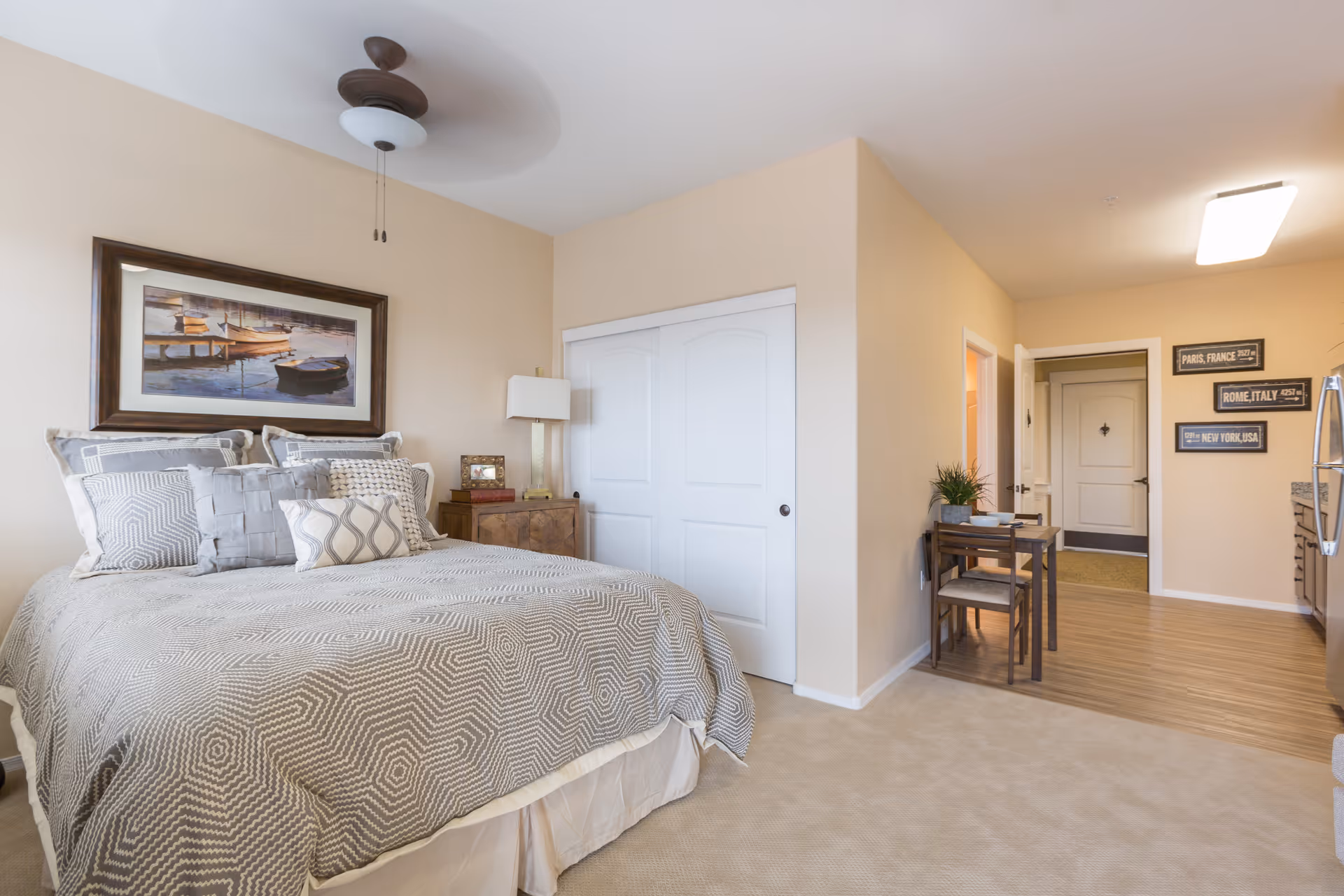 A cozy bedroom in a senior living facility with a neatly made bed featuring patterned bedding and multiple pillows. Above the bed is a framed picture of boats on water. To the right of the bed is a wooden nightstand with a lamp and decorative items. The room has beige walls and carpeted flooring. Adjacent to the bedroom area is a small dining table with two chairs, and a hallway leading to other rooms. The space is well-lit with a ceiling fan and overhead light.