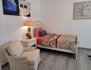 A cozy bedroom with a single bed featuring patterned bedding and pillows. Next to the bed is a small white nightstand with a lamp and a small flower arrangement. A comfortable beige armchair is positioned near the bed. The room has light-colored walls with two framed paintings hanging on them and a gray wooden floor.