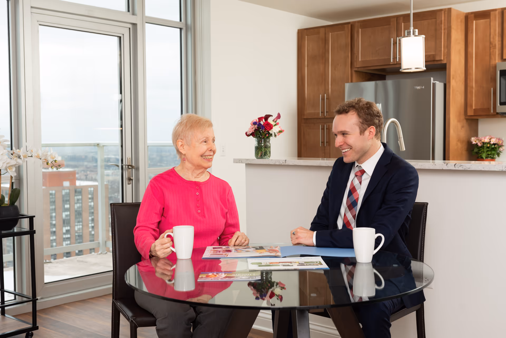 An elderly woman in a bright pink sweater and a man in a suit with a plaid tie sit at a round glass table in a modern kitchen area. They are smiling and appear to be engaged in a pleasant conversation. The table has two white mugs and some brochures or magazines on it. Behind them is a kitchen counter with wooden cabinets, a stainless steel refrigerator, and a vase with flowers. Large windows and a glass door reveal a cityscape outside.