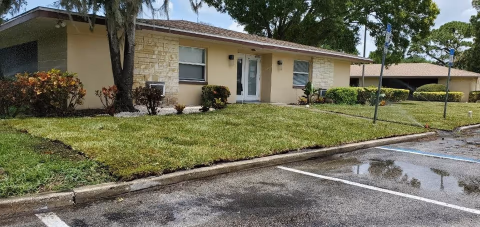 Exterior view of a single-story assisted living facility building with beige walls, stone accents, and a brown shingle roof. The building is surrounded by a well-maintained lawn, bushes, and trees. In front of the building, there is a parking lot with marked parking spaces, including handicapped spots.