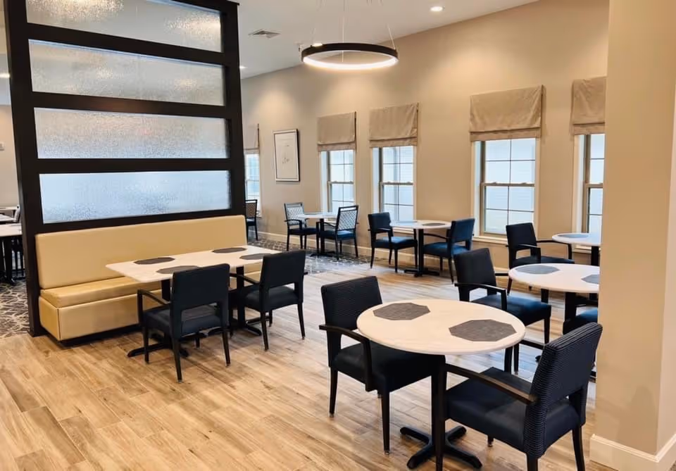 A bright dining room with multiple tables and chairs arranged neatly. The room features light wood flooring, beige walls, and several windows with beige roman shades. There is a cushioned bench along a partition wall, and modern circular light fixtures hang from the ceiling.