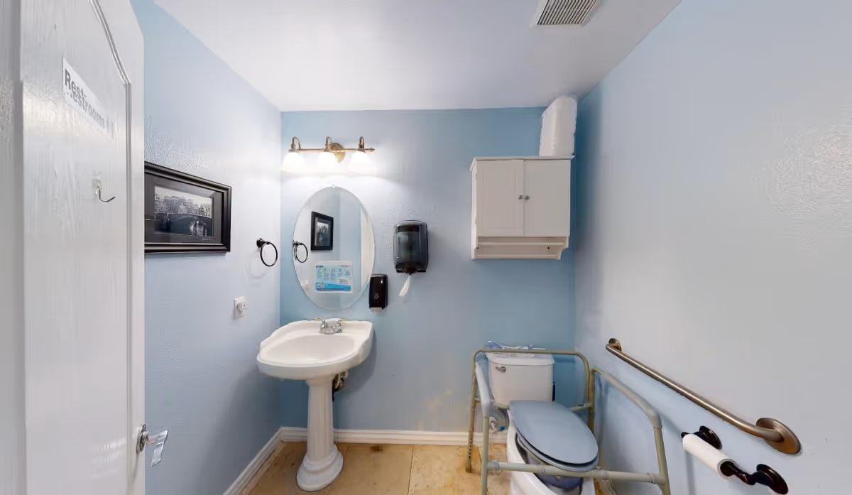 A small bathroom with light blue walls featuring a white pedestal sink with an oval mirror above it, a white cabinet mounted on the wall, a toilet with safety rails around it, a handrail on the wall, and a door labeled 'Restrooms'.