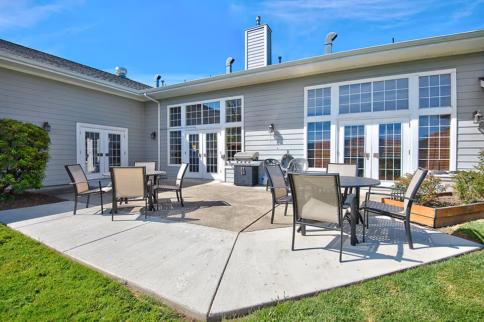 Outdoor patio area at Bridgewood Rivers Assisted Living featuring multiple round tables with chairs arranged around them on a concrete surface. The patio is adjacent to a building with large windows and glass doors, under a clear blue sky. There is a grill and some plants in raised garden beds nearby.