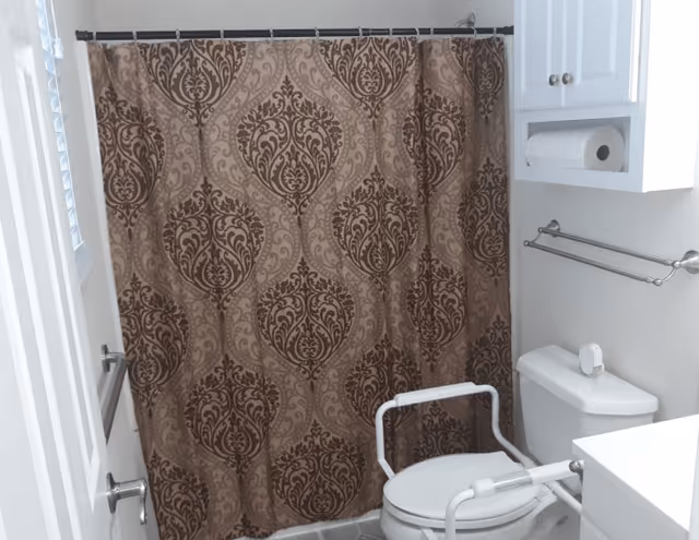 Bathroom with a patterned brown shower curtain, a white toilet with safety rails, a white cabinet with a roll of toilet paper, and a towel rack on the wall.