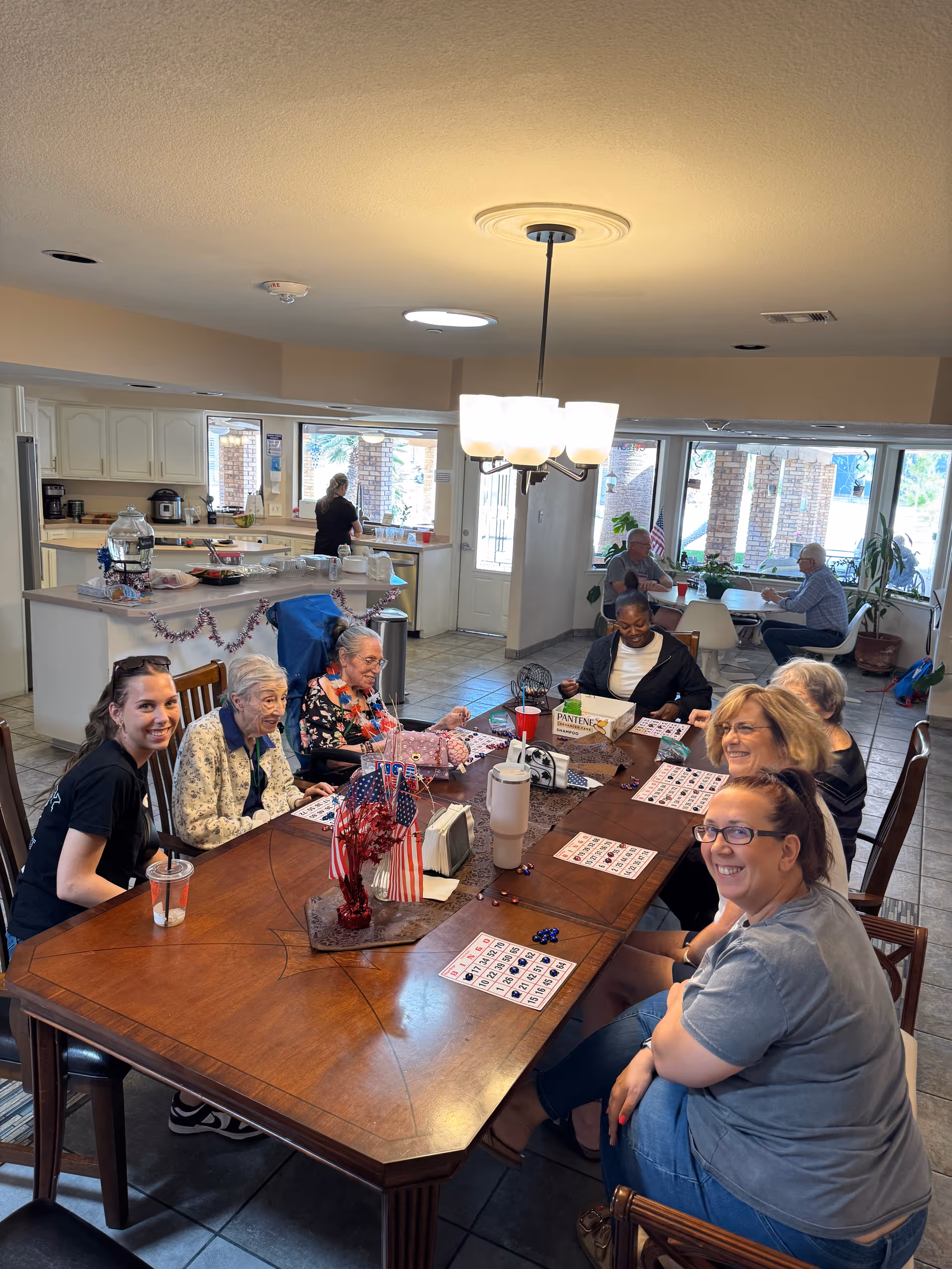 A group of seniors and caregivers gathered around a long wooden table in a communal dining/kitchen area playing bingo.