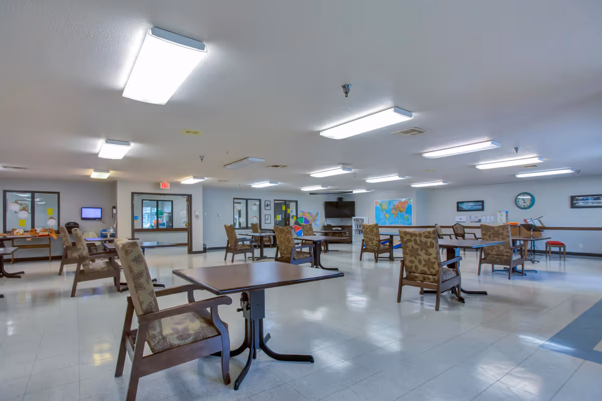 A spacious, well-lit common area with multiple tables and chairs arranged throughout the room. The walls are decorated with a world map, a clock, and framed pictures. There is a television mounted on the far wall and a bookshelf underneath it. The floor is tiled, and the ceiling has several fluorescent light fixtures.