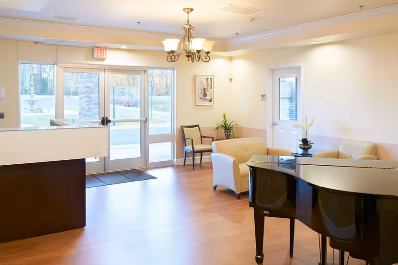 Bright and welcoming assisted living facility lobby with wooden flooring, a black grand piano, cream-colored armchairs, a small table with a flower vase, a chandelier, and glass double doors leading outside.