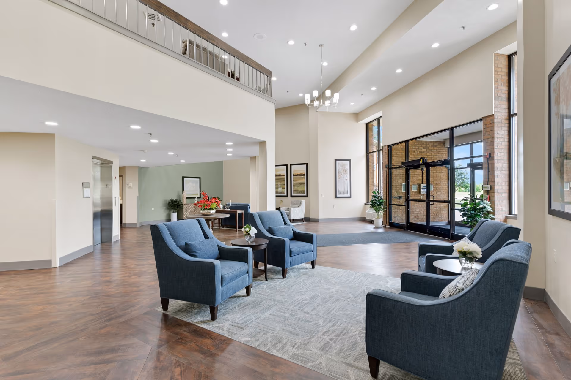 Spacious and well-lit senior living facility lobby with four blue armchairs arranged around two small round tables on a patterned rug. Large windows and glass doors allow natural light to fill the space. The lobby features wood flooring, a high ceiling with recessed lighting and a modern chandelier, framed artwork on the walls, and a reception desk with flowers in the background.