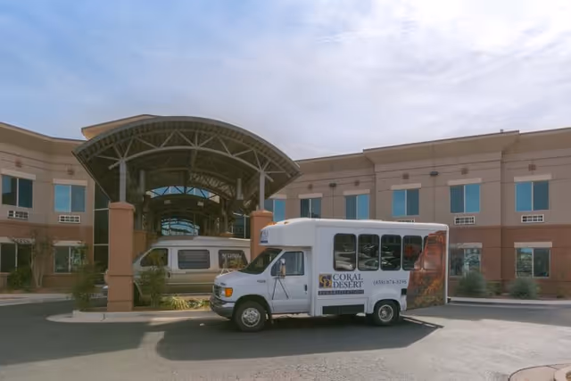 Exterior view of Coral Desert Rehabilitation And Care facility showing a two-story building with multiple windows and a covered entrance. A white shuttle bus with the Coral Desert logo is parked in front of the entrance under a clear sky.