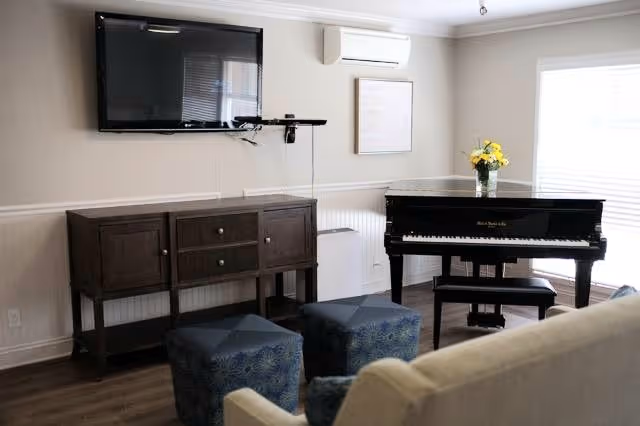 Sunny common room with a black upright piano, wall-mounted TV, wooden console table, blue ottomans and seating with a vase of yellow flowers.