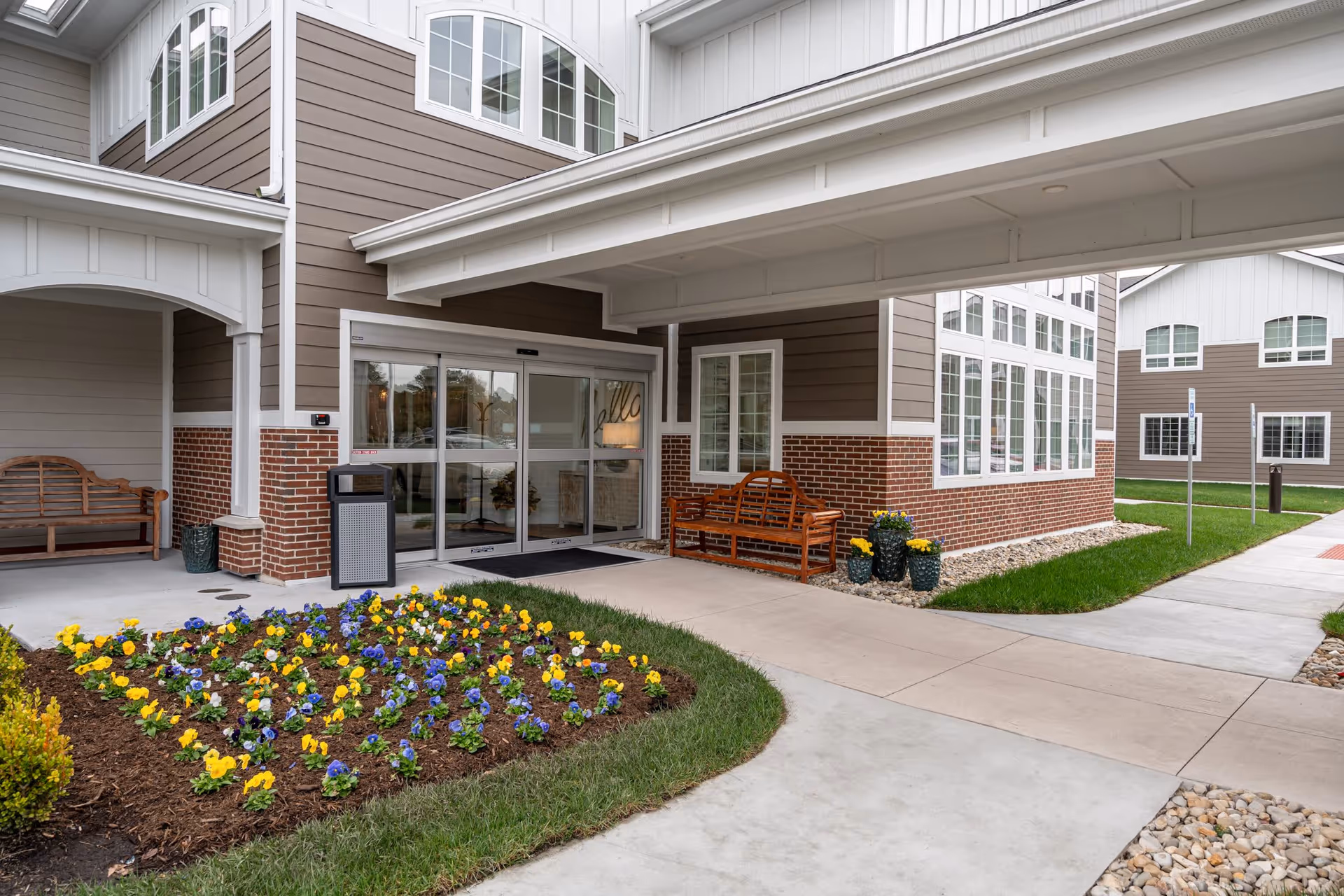 Entrance to a senior living facility with sliding glass doors, two wooden benches on either side, a flower bed with yellow and purple flowers, and a covered walkway. The building exterior features brown siding with white trim and brick accents.