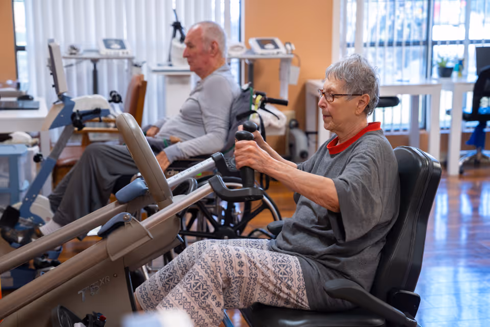 Two elderly individuals exercising on seated fitness machines in a well-lit room with wooden floors and large windows. One person is in the foreground using a rowing machine, while the other is in the background on a similar exercise device.