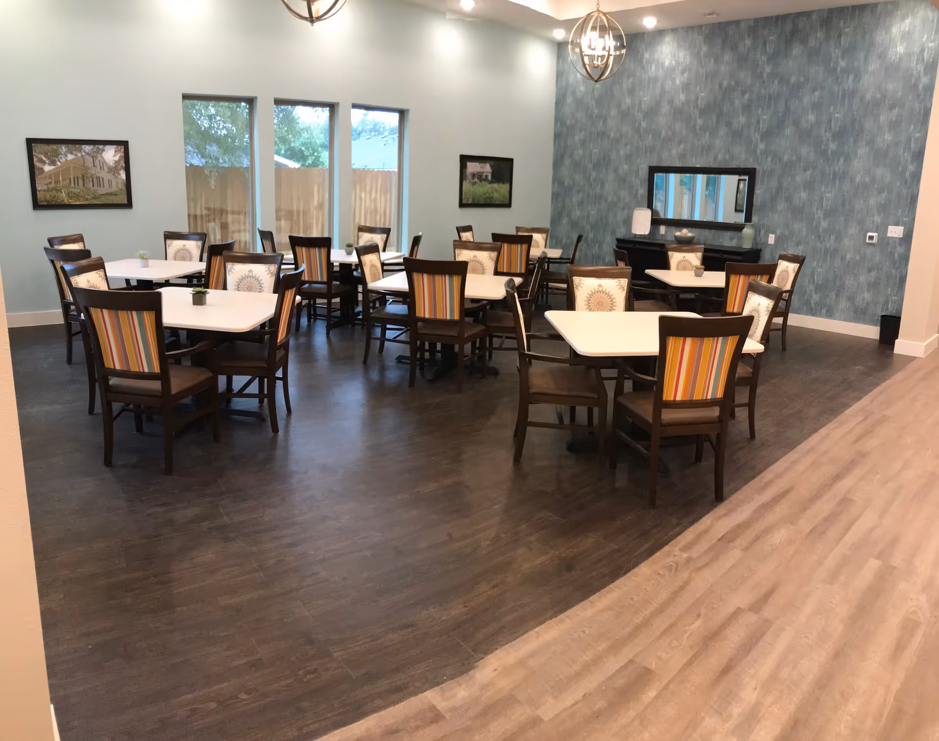 A dining room with several square tables and wooden chairs with striped and patterned cushions. The room has large windows with a view of a wooden fence outside, light blue walls, two framed pictures, and two modern pendant lights hanging from the ceiling. The floor is a combination of dark and light wood tones.