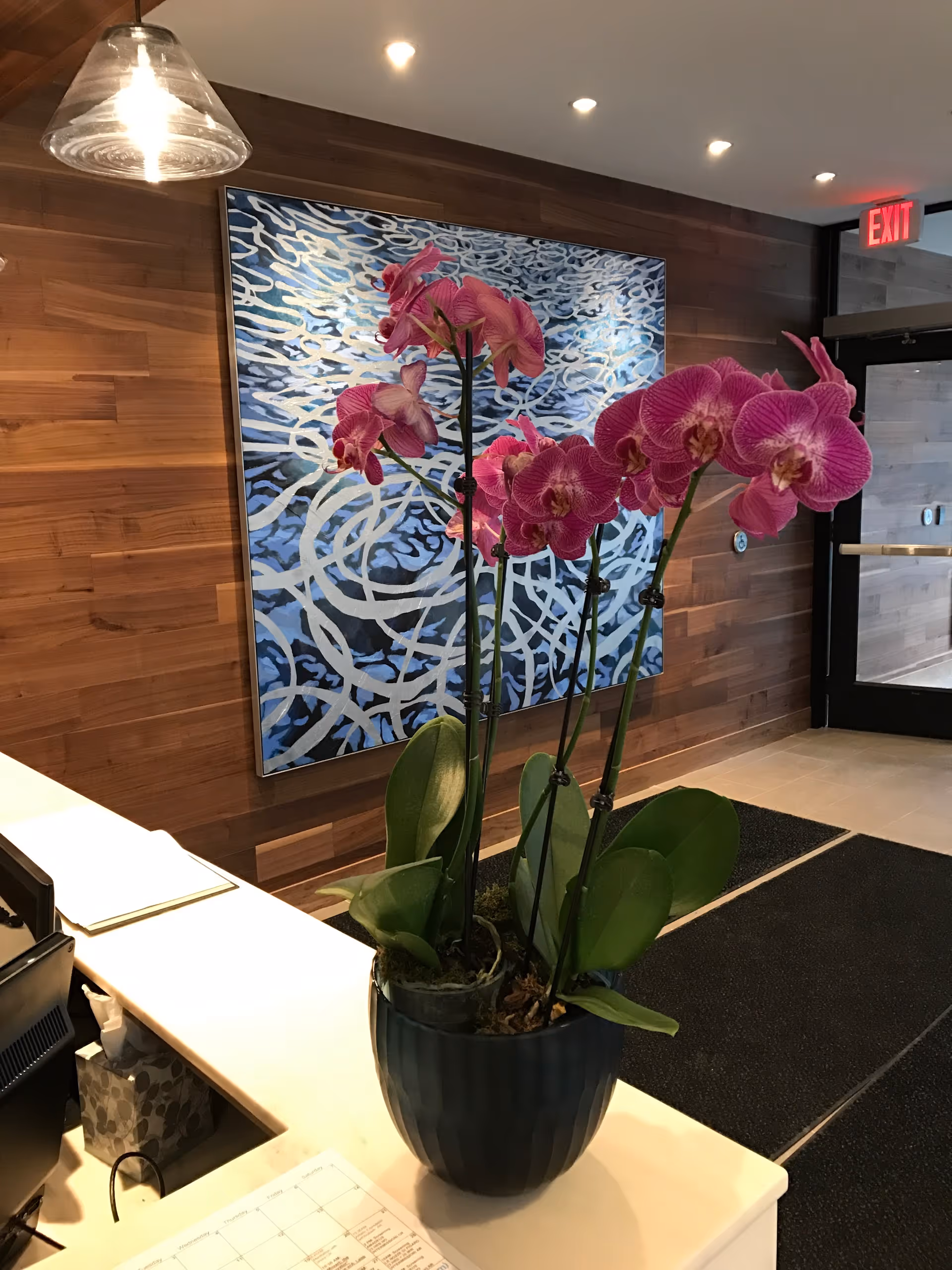 Potted pink orchids on a reception desk in a wood-paneled lobby with abstract artwork and an exit door.
