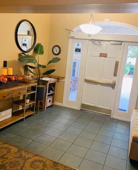 Foyer of an assisted living facility with a front double-door entrance, tiled floor, console table with decor, a large potted plant and wall mirror.