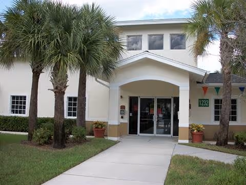 Entrance to a building with a covered walkway supported by columns, flanked by palm trees and potted plants. The building is light-colored with a sign displaying the number 1232 near the door.