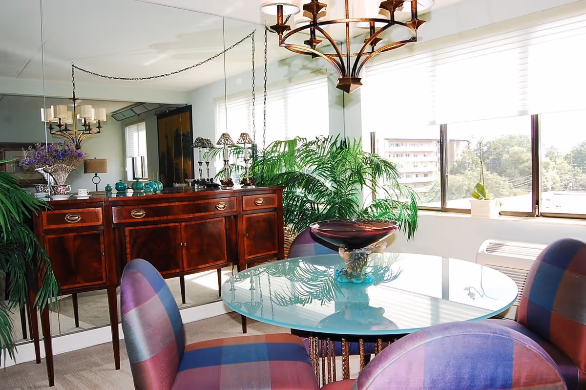 A bright dining area with a round glass table surrounded by four colorful plaid upholstered chairs. Behind the table is a wooden sideboard with decorative items including a vase with purple flowers and teal ceramic pieces. Large green plants are placed near the window, which lets in natural light. A chandelier with multiple candle-style lights hangs above the table. The wall behind the sideboard is covered with a large mirror reflecting the room.