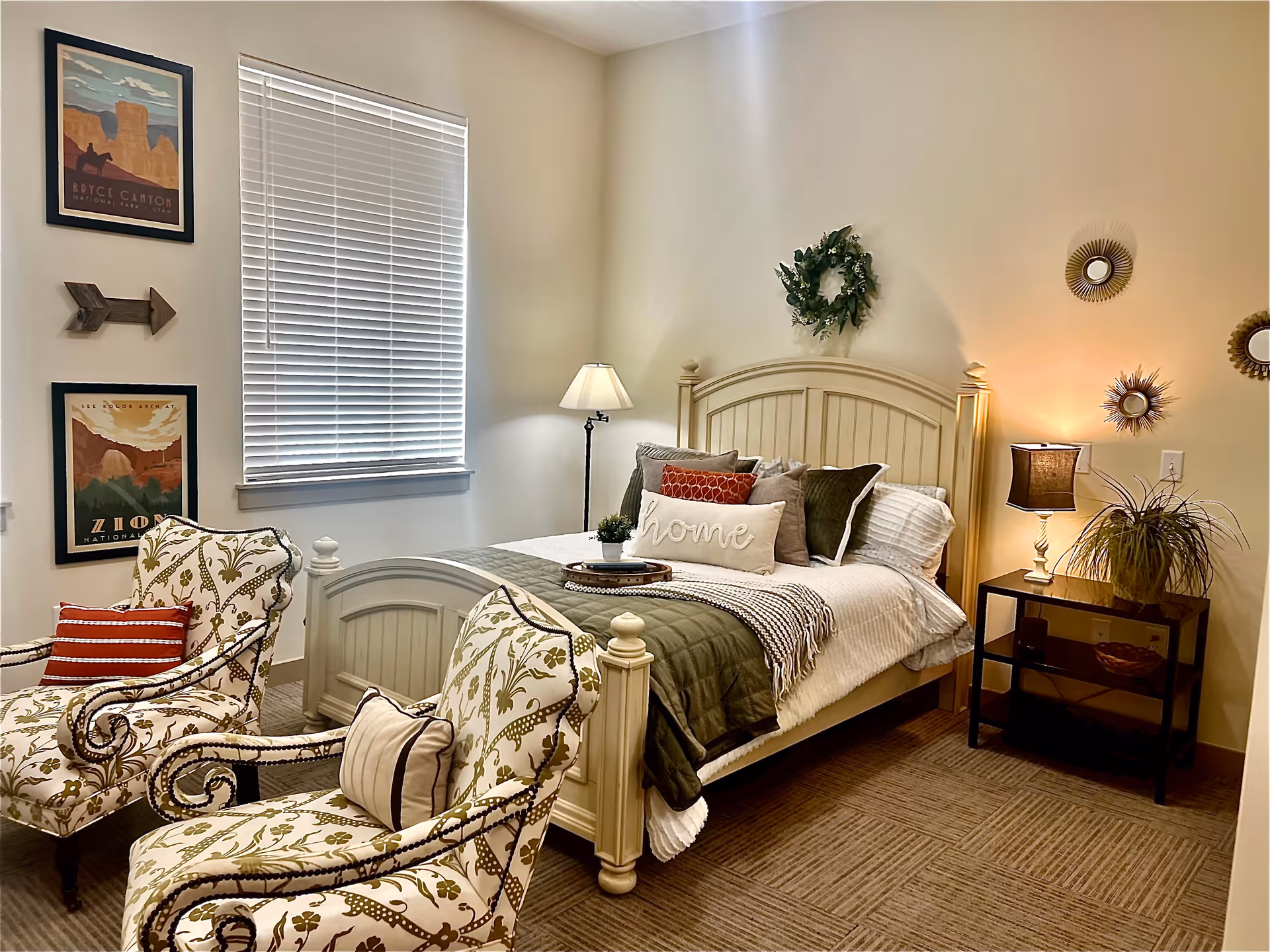 A cozy bedroom with a cream-colored wooden bed adorned with multiple pillows and a throw blanket. Two patterned armchairs with cushions are placed near the foot of the bed. A window with closed blinds is on the left wall, flanked by two framed posters and a wooden arrow decoration. A floor lamp stands beside the bed, and a nightstand with a table lamp and a potted plant is on the right. Decorative wreath and sunburst mirrors hang on the walls.