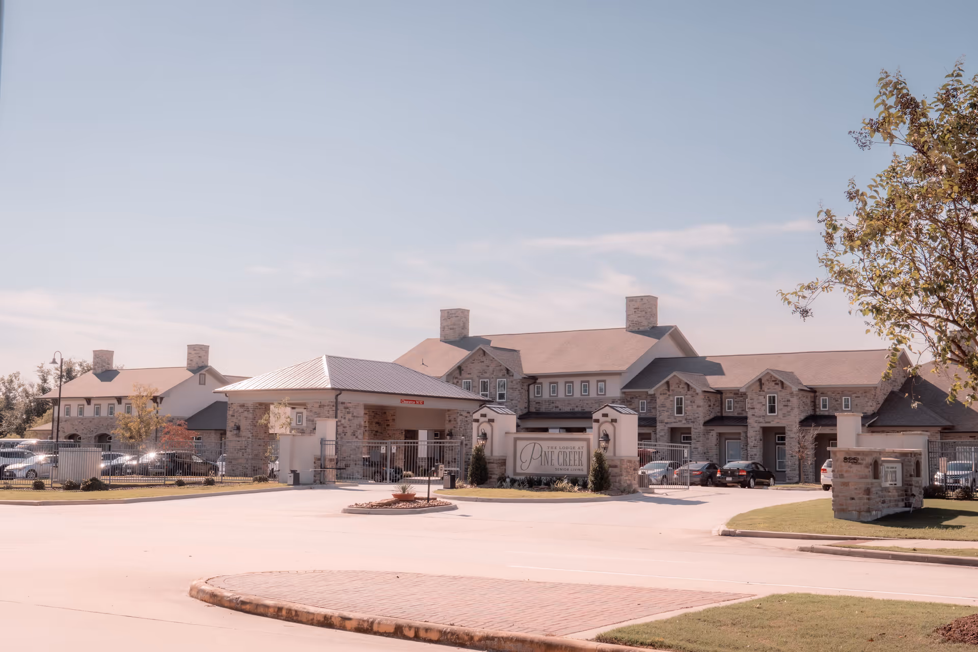 Exterior view of The Lodge at Pine Creek, a large stone and brick senior living facility with multiple chimneys, a covered entrance, and a parking area with several cars. The sky is clear with some light clouds, and there is a tree on the right side of the image.