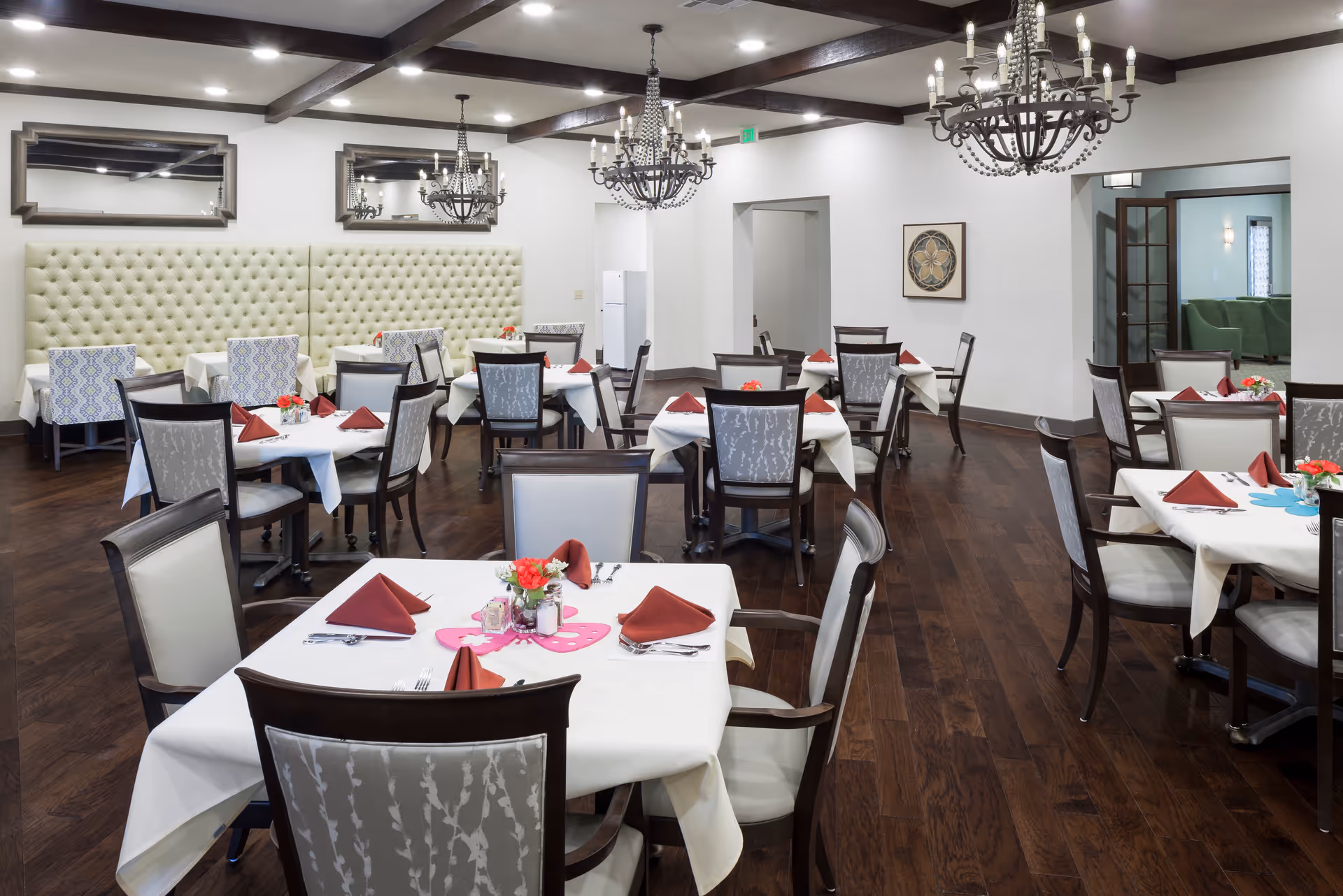 A dining room with multiple tables covered in white tablecloths, each set with red folded napkins, silverware, and small flower centerpieces. The room features dark wood flooring, tufted beige wall padding, decorative mirrors, and ornate chandeliers hanging from a ceiling with exposed wooden beams.