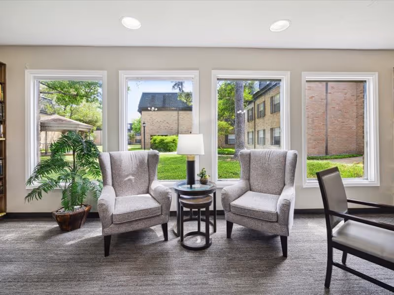 A bright sitting area with two gray upholstered armchairs facing each other with a small round table and lamp between them. There is a potted plant to the left and large windows behind the chairs showing a green outdoor area with trees and buildings.