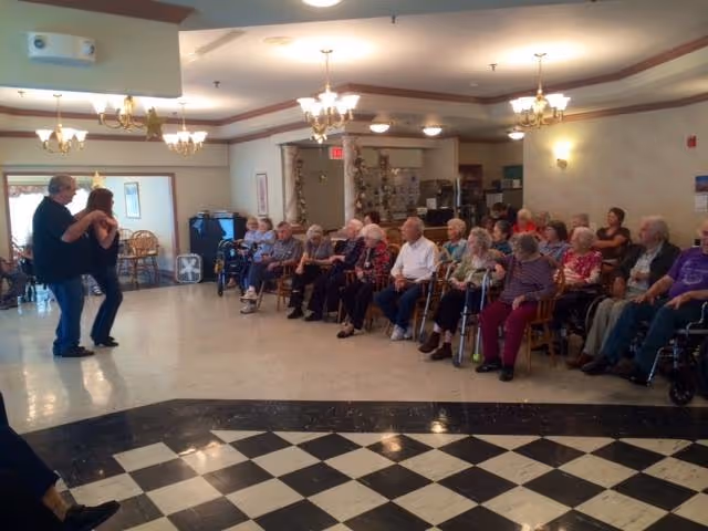 A group of elderly people seated in chairs along the wall of a large room, watching two people dancing on a checkered floor area. The room has chandeliers and a light-colored interior with some furniture and decorations.