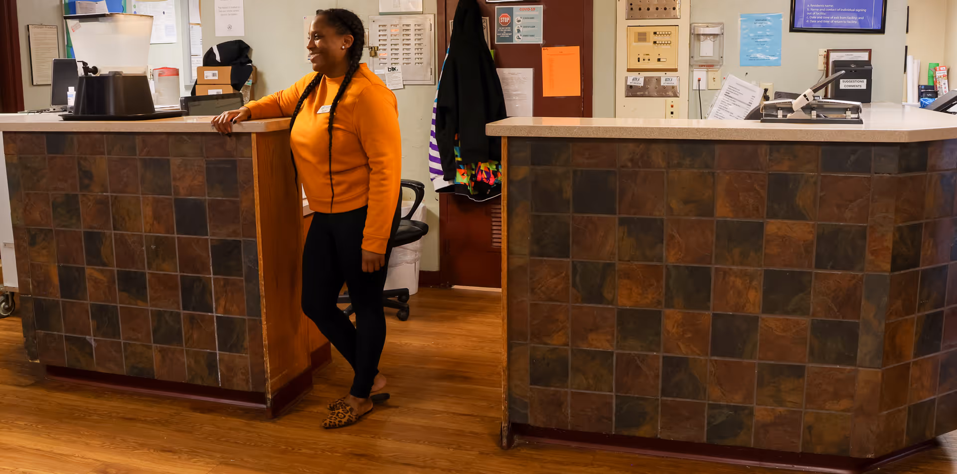 A woman in an orange sweater and black pants stands and leans against a tiled reception desk in a facility office area with wood flooring and various office supplies and notices on the walls behind her.