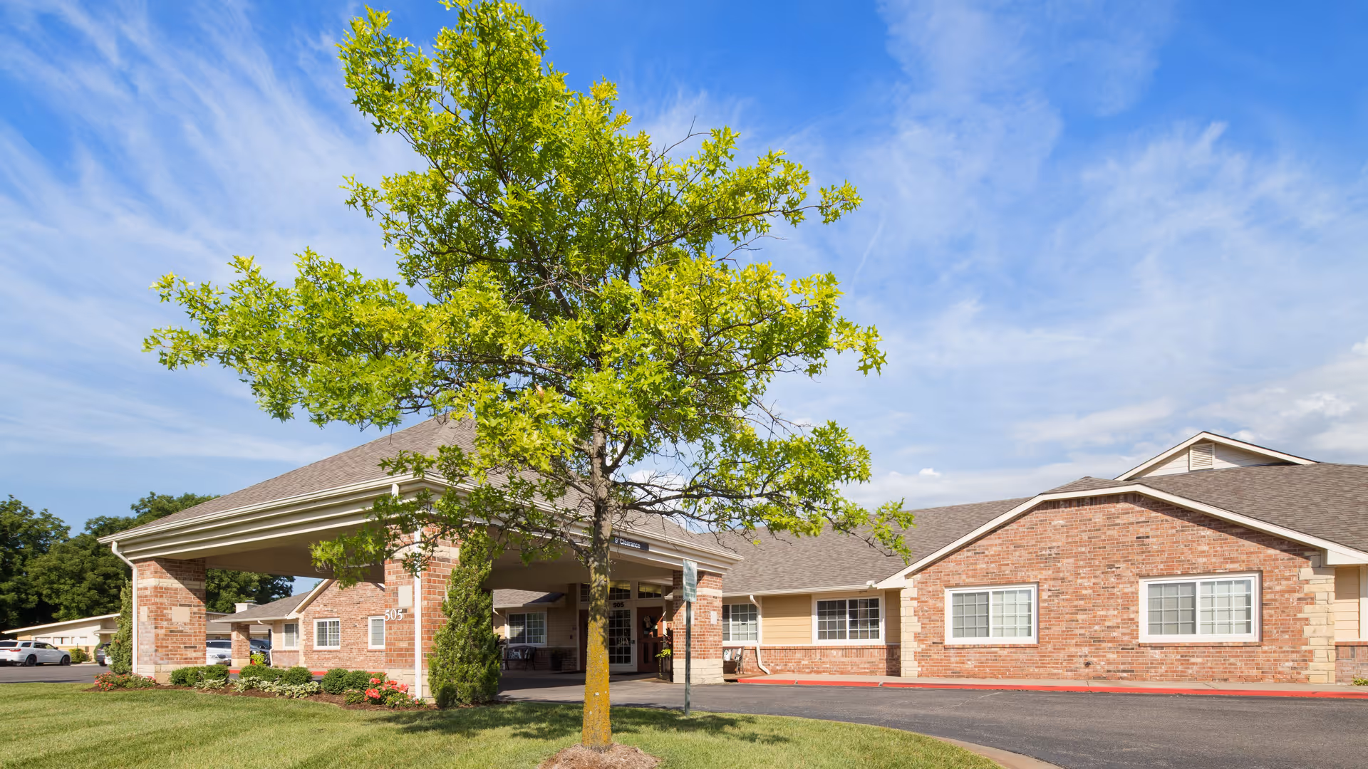 Exterior view of Park West Plaza facility showing a brick building with a covered entrance, a tree with green leaves in the foreground, a clear blue sky with some clouds, and a parking area with cars.
