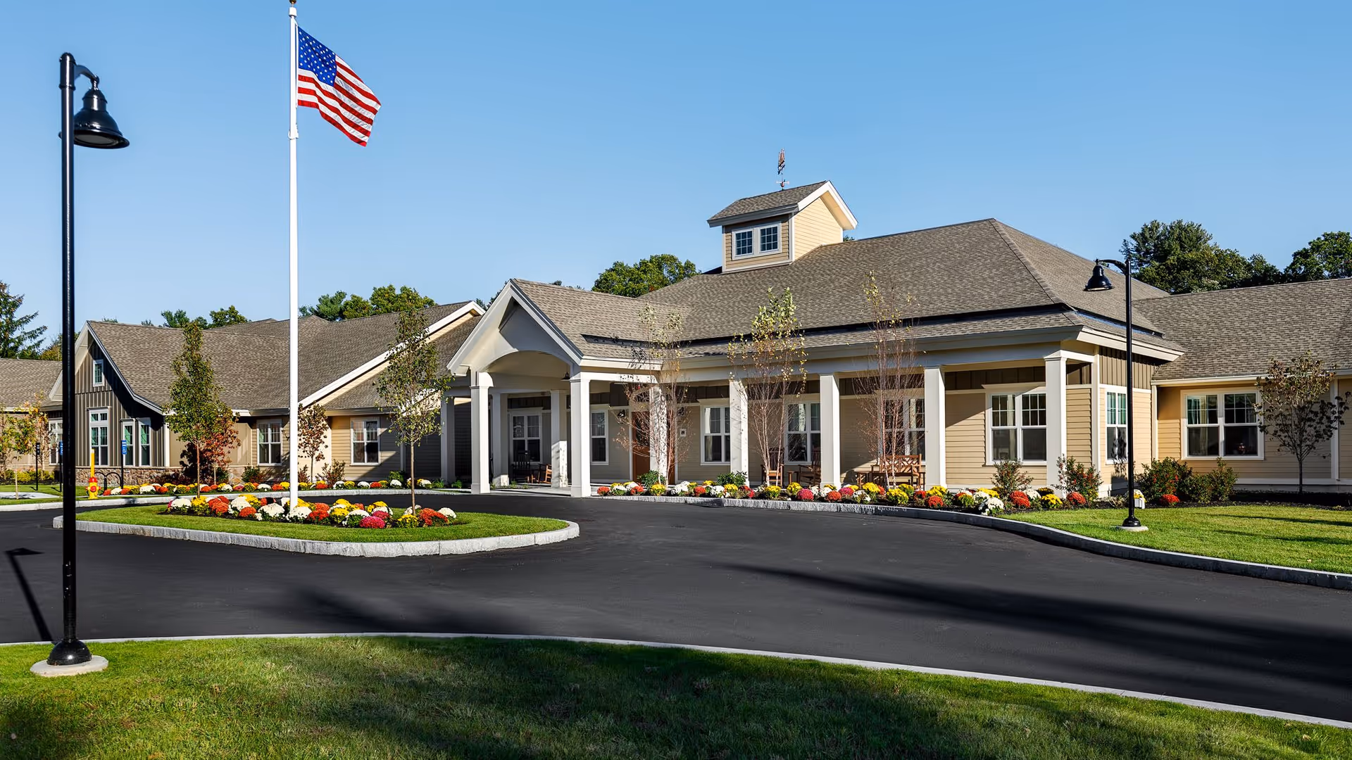 Front entrance of a single-story senior living facility with a circular driveway, an American flag, and landscaped flower beds.