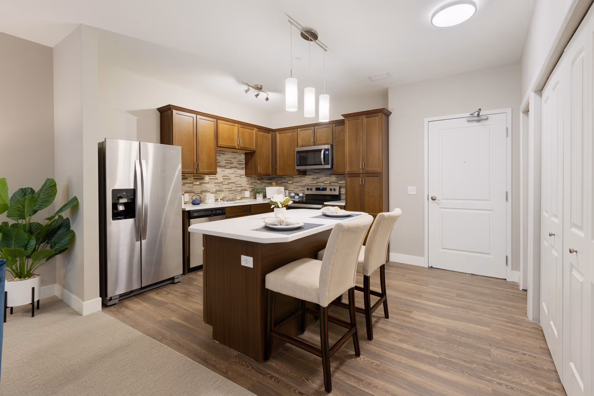 Modern kitchen with wooden cabinets, stainless steel refrigerator, microwave, and stove. A kitchen island with a white countertop has two beige upholstered chairs and place settings. The floor is wood, and there is a potted plant in the corner near the carpeted area.