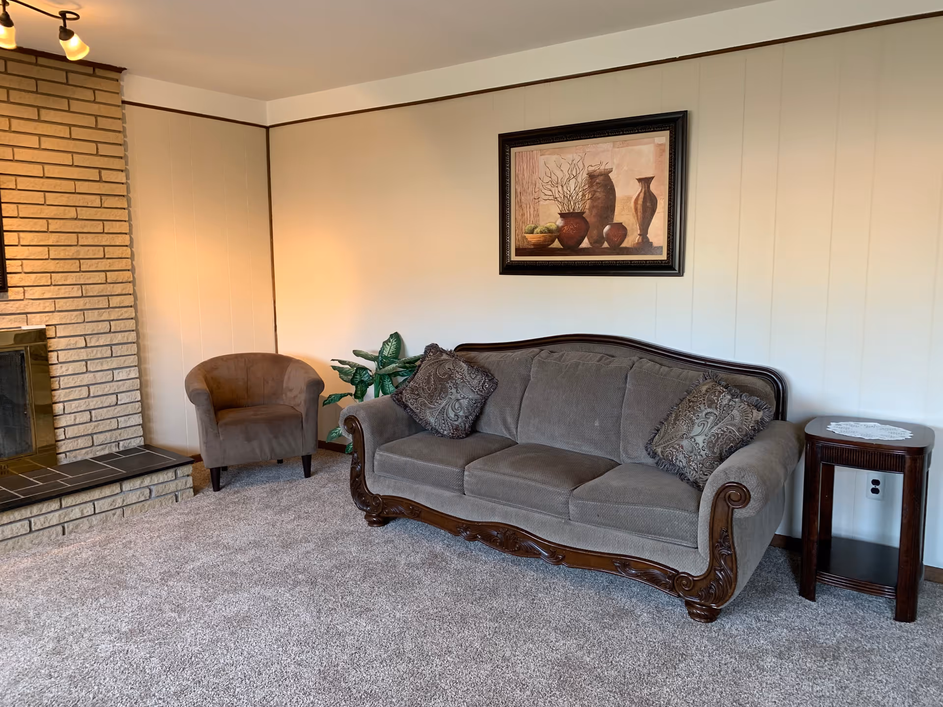 A cozy living room with a gray upholstered sofa, patterned throw pillows, an armchair, a brick fireplace, and framed wall art.