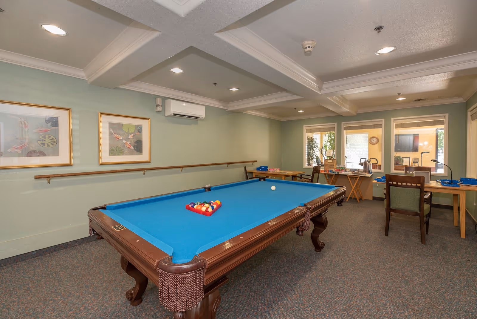 Interior room with a blue felt pool table in the center, two framed pictures on the wall, several tables and chairs near windows, and a ceiling with recessed lighting.