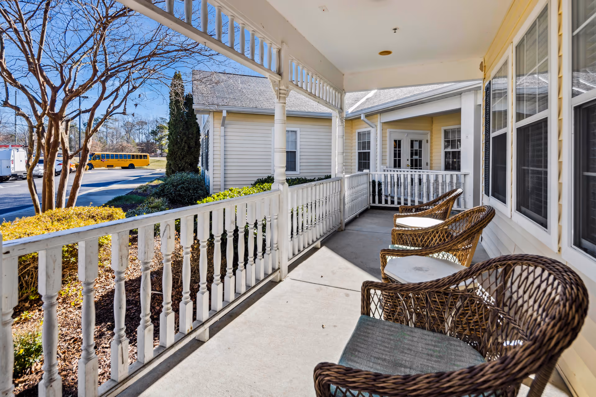 Covered front porch with wicker chairs and white railing at a senior living facility, with a yellow school bus visible in the parking area beyond.