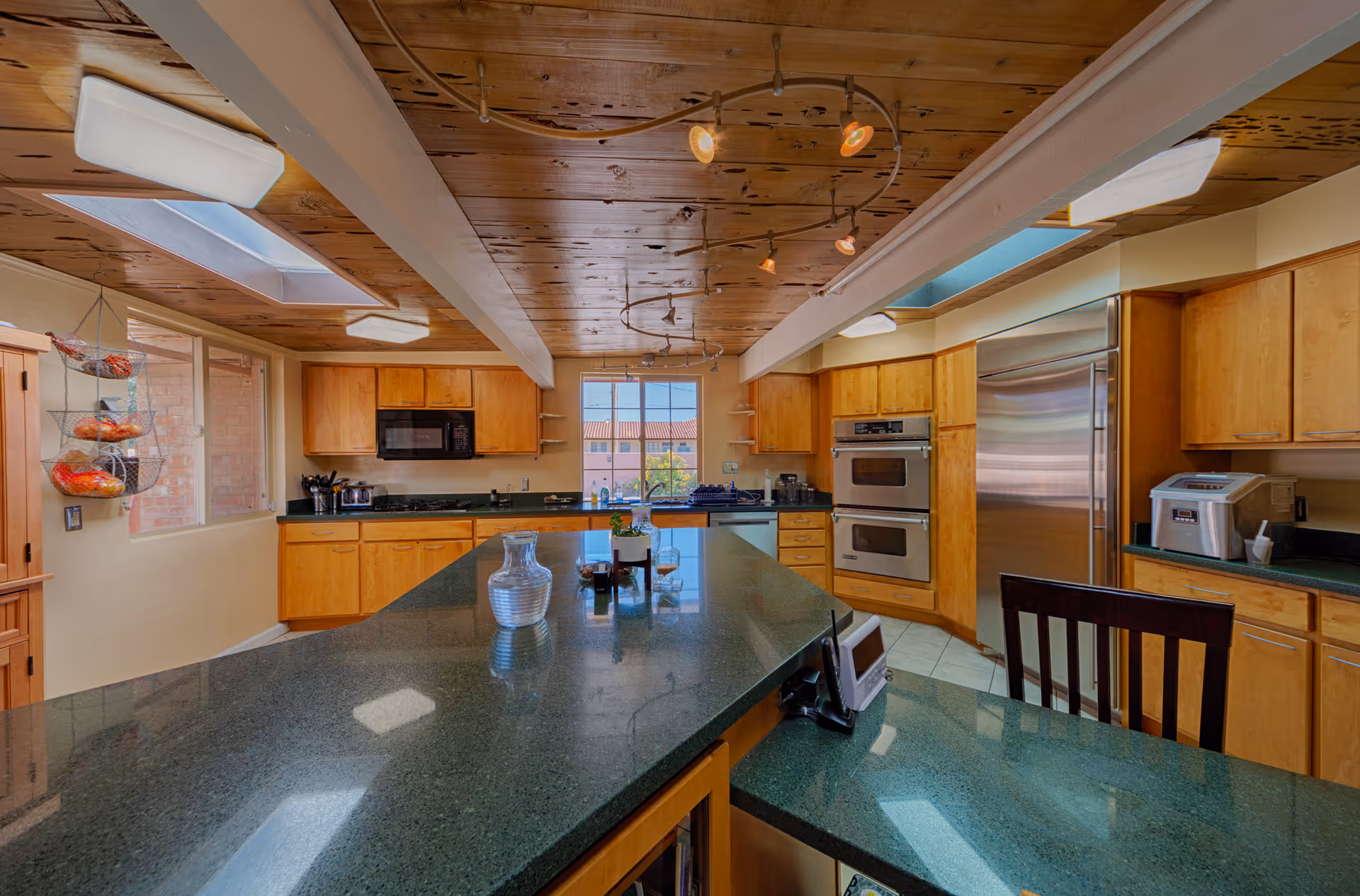 Spacious kitchen with wooden cabinets, a large green granite island countertop, stainless steel double oven, refrigerator, microwave, and various kitchen appliances. The ceiling is wooden with exposed beams and modern track lighting. There are windows and skylights allowing natural light to brighten the room.