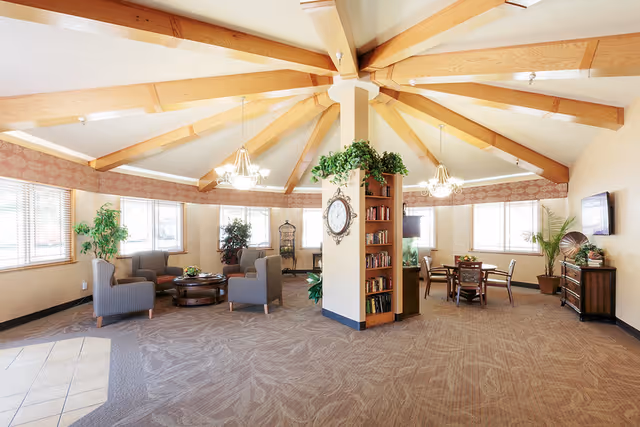 A spacious, well-lit common area with a high ceiling featuring exposed wooden beams radiating from a central column. The room is furnished with comfortable armchairs arranged around a circular coffee table on the left side and a dining table with chairs on the right. The central column has a clock and built-in bookshelves with plants on top. Large windows allow natural light to fill the room, and additional plants and decorative items are placed around the space.
