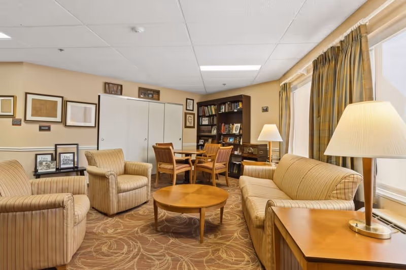 A cozy living room area with beige striped armchairs and a matching sofa surrounding a round wooden coffee table. There is a wooden side table with a lamp on the right side and a bookshelf filled with books in the background. The walls are decorated with framed pictures and the room has large windows with patterned curtains letting in natural light.
