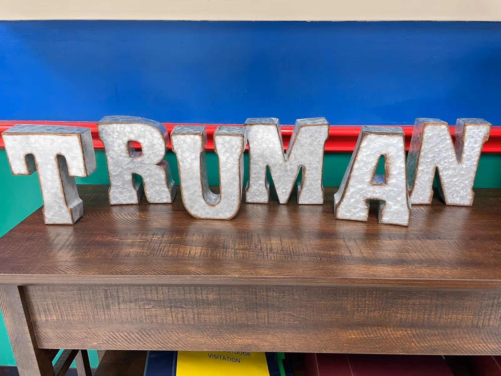 Metal decorative letters spelling TRUMAN arranged on a wooden table against a blue and green wall.