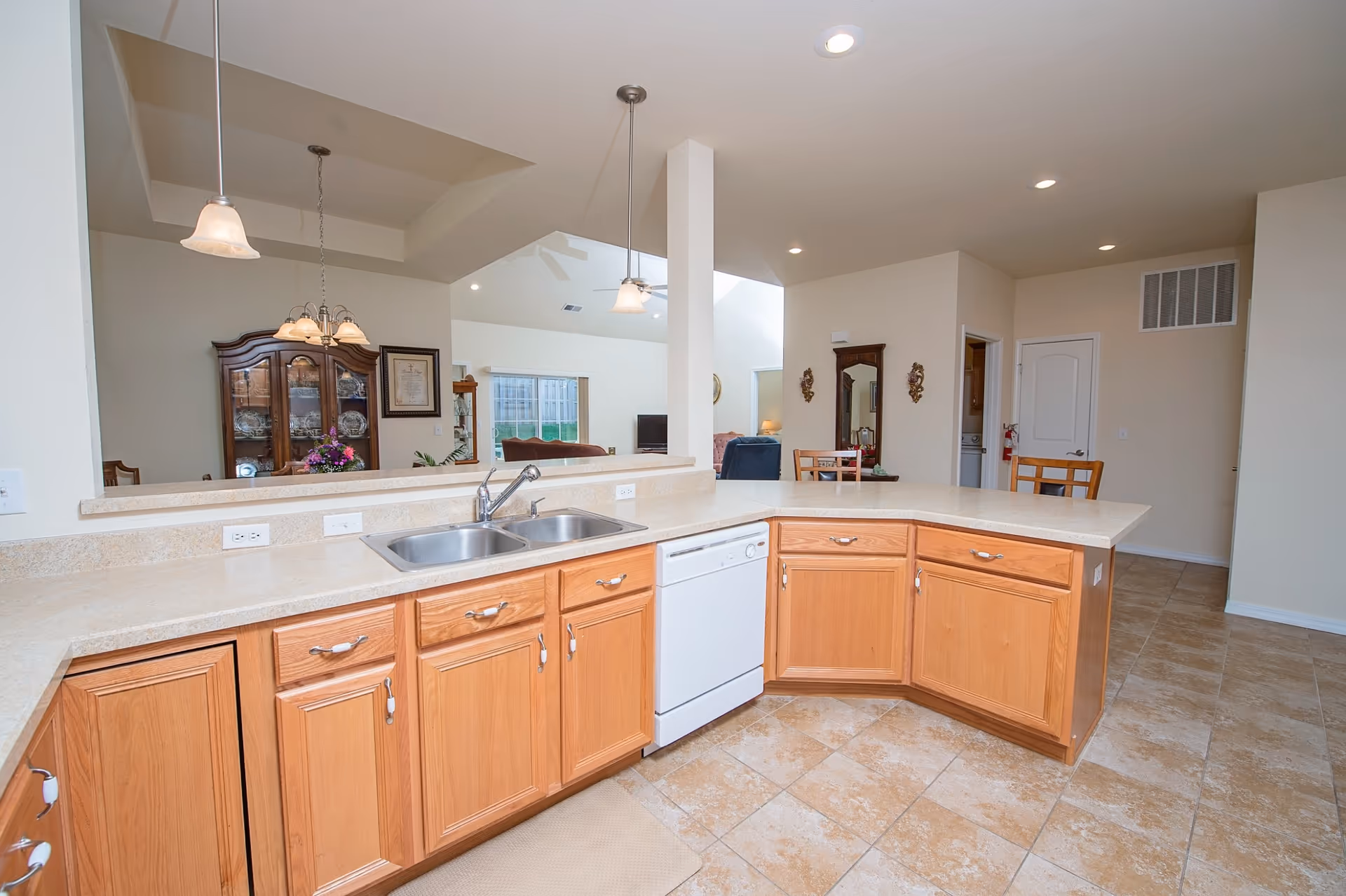 A spacious kitchen area with light wood cabinets, a double sink, and a white dishwasher. The kitchen counter extends into a breakfast bar with two pendant lights hanging above. In the background, there is a dining area with a wooden china cabinet, a dining table with chairs, and a living room with a TV and large windows letting in natural light.